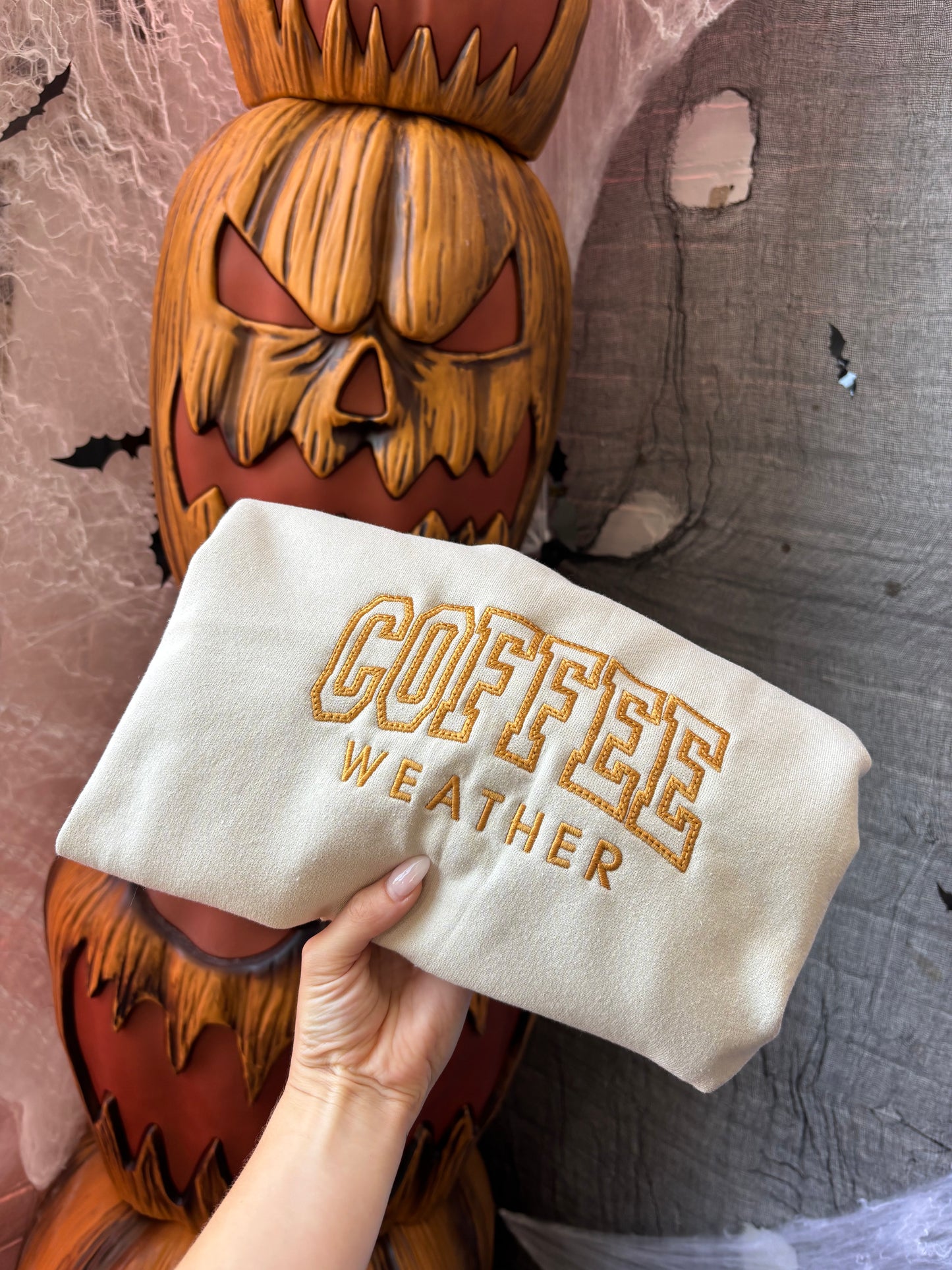 Hand holding a pouch with 'Coffee Weather' text in front of a carved pumpkin decoration.