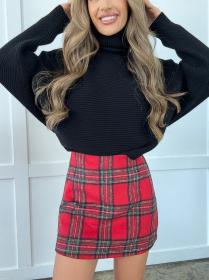 Person wearing a black sweater and red plaid skirt against a white background