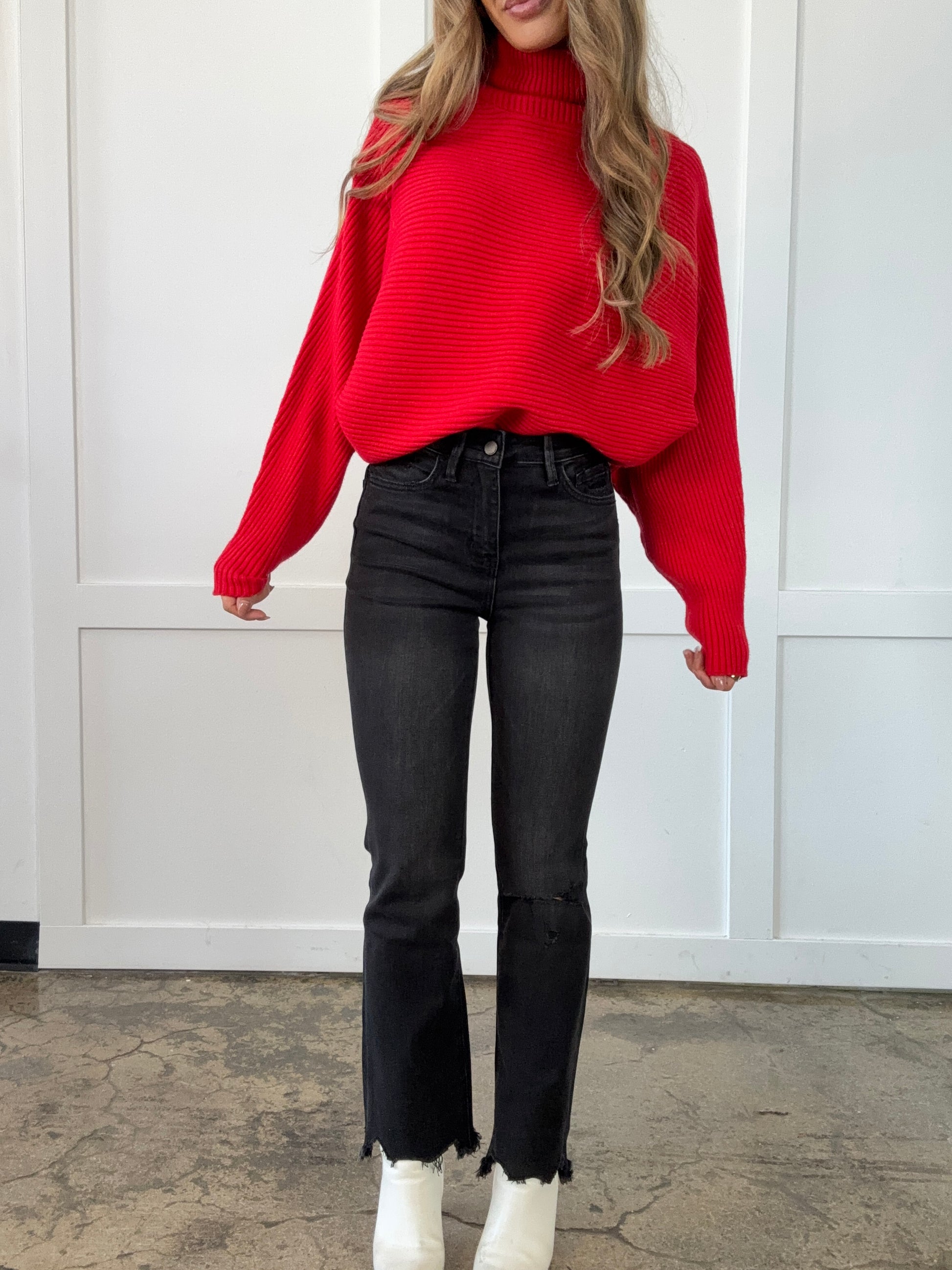 Person wearing a red sweater and black pants standing against a white door.