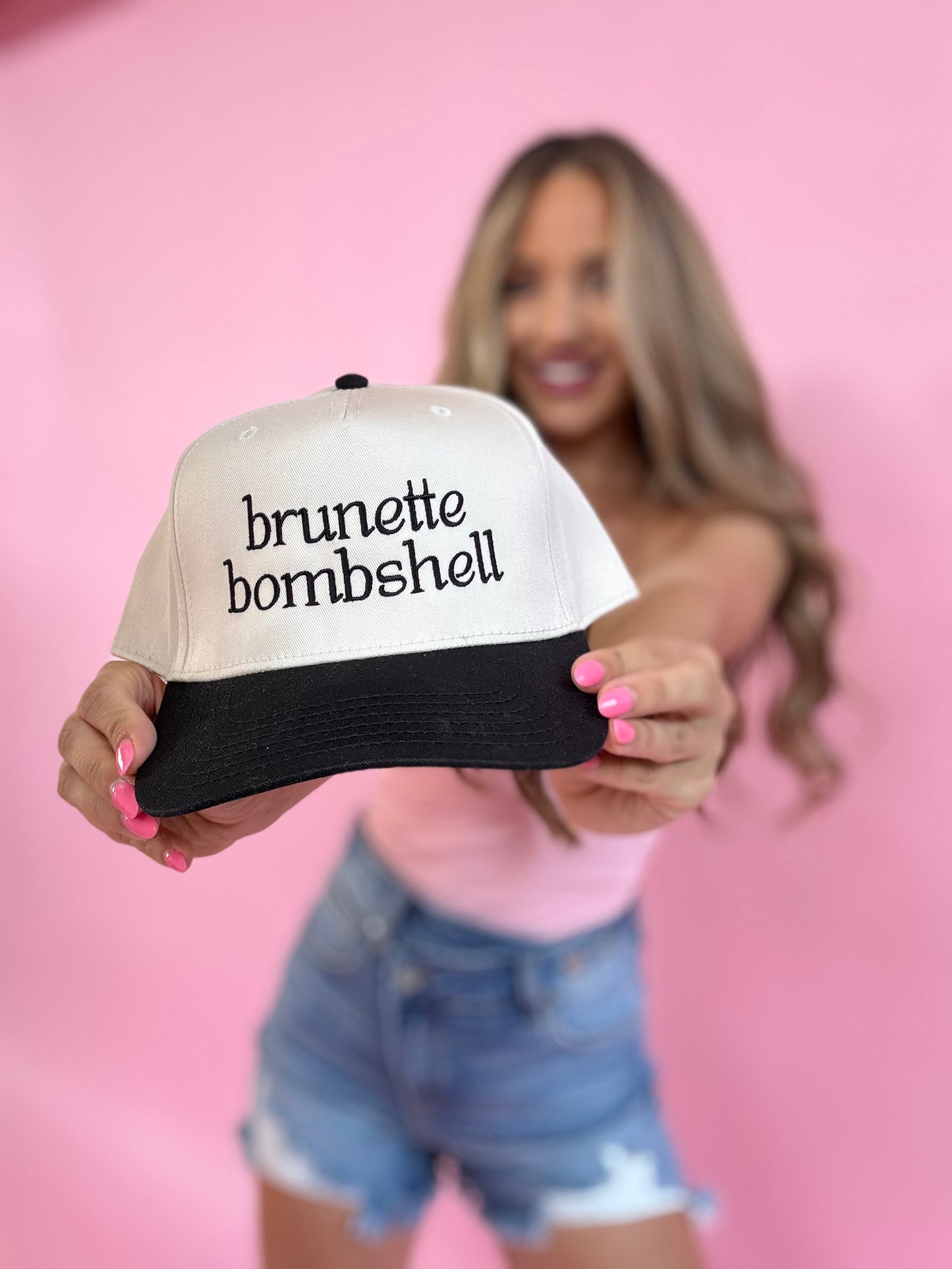 Woman holding a cap with 'brunette bombshell' text against a pink background
